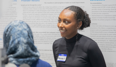 A student in front of her research poster with a name tag smiling at a woman in front of her.