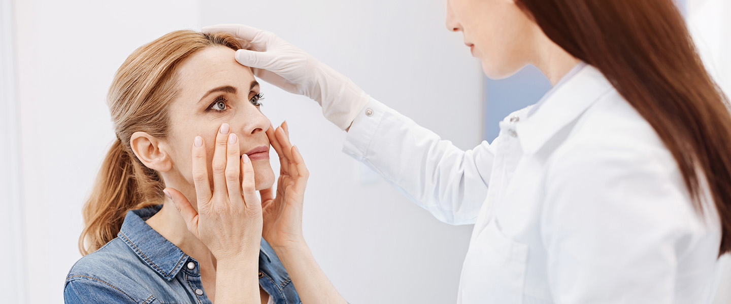 A concerned woman touches her face while being examined by a doctor.