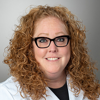 Carrie Dudek has long curly red hair, is wearing black frame glasses, a black shirt, and white medical coat. She is smiling at the camera.