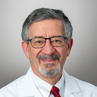Dr. Kanaan has short gray hair, gray goatee and mustache, is wearing glasses, a white dress shirt and red tie under a white medical coat, and is smiling at the camera