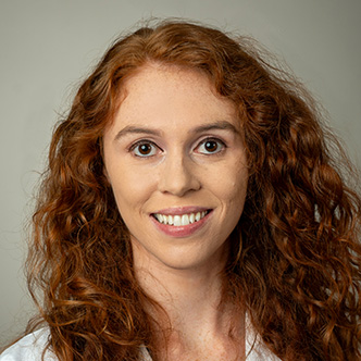 Daniella Schneider has long curly red hair, is wearing a white medical coat and is smiling at the camera.