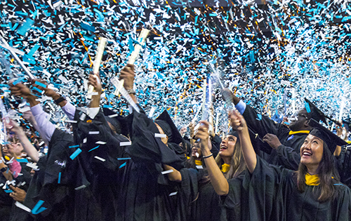As blue confetti falls, graduates in caps and gowns wave their diplomas in the air