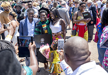 A gowned Black female graduate poses for a photograph with two family members in a crowd of celebrating grads and family members. 