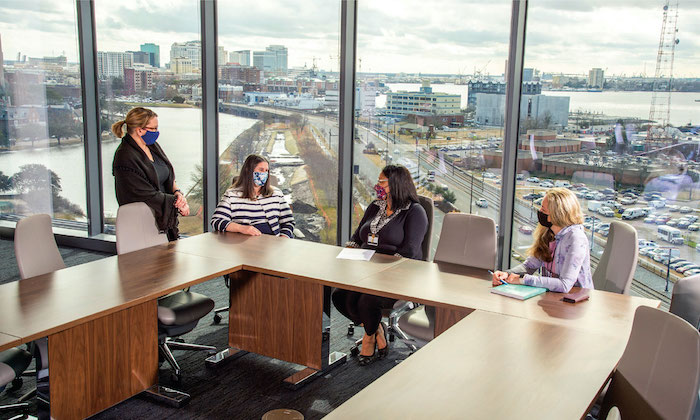 Co-workers meet in a conference room on a top floor of Waitzer Hall. A dramatic view of downtown Norfolk and the surrounding waterfront is visible behind them.
