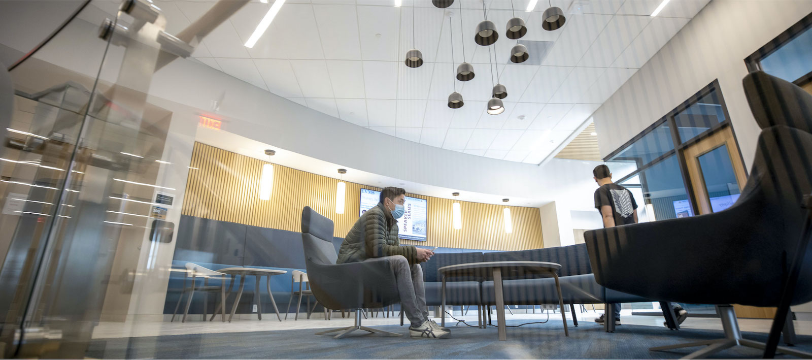 One of several lobby areas in Waitzer Hall furnished with modern tables and chairs; and outfitted with wood paneling, wall sconces and overhead light fixtures. Lobby areas in the education podium provide space for students to mix (post-COVID-19), study or relax.