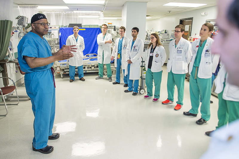 Clad in blue surgical scrubs, Dr Britt stands in a hospital hallway talking to large group of residents.