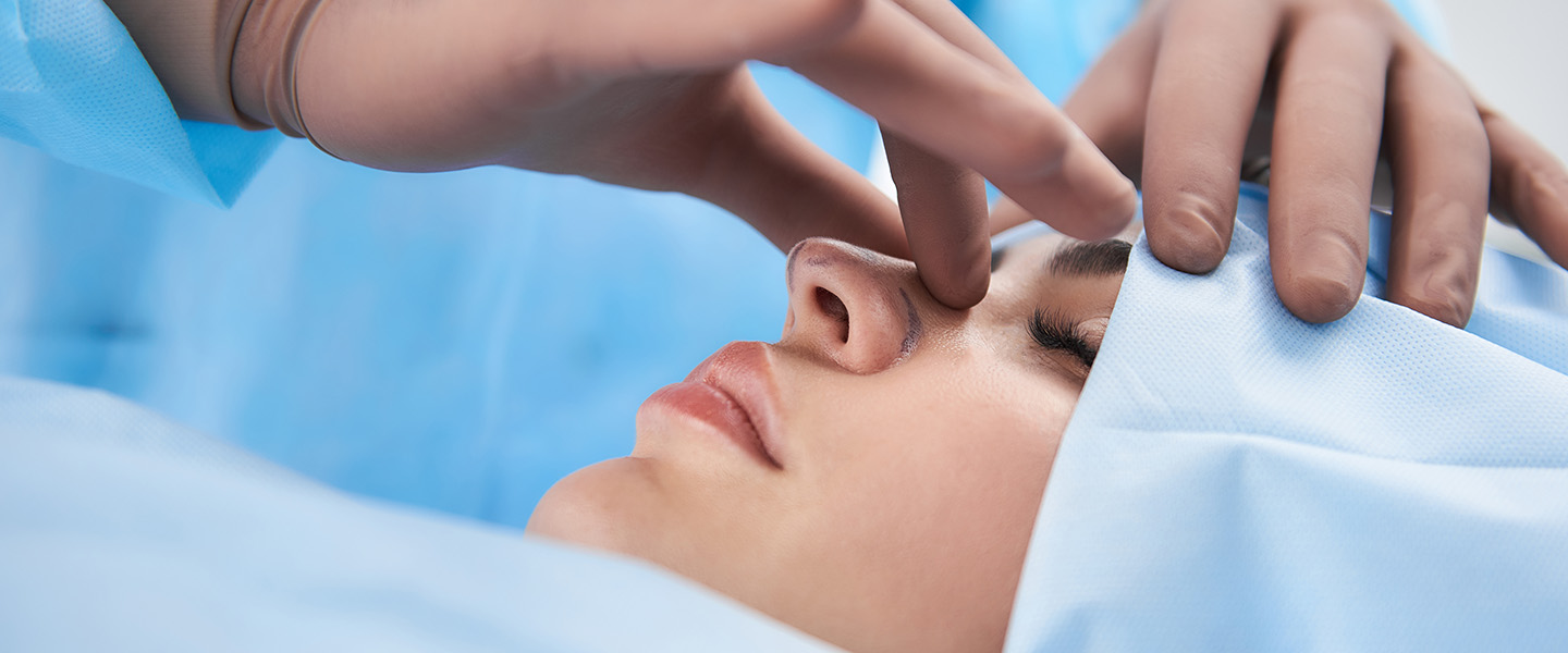 A woman wearing blue scrubs and hair cover is undergoing a rhinoplasty.