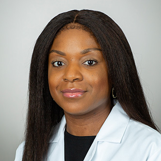 Joycelyn has long dark hair, is wearing a white medical coat and dark shirt, and is smiling at the camera