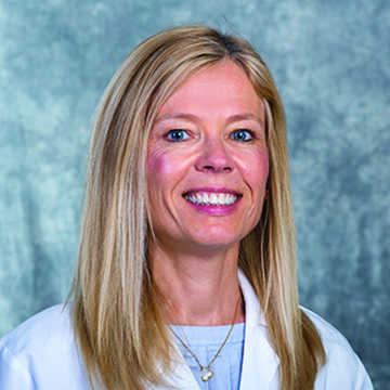 Dr. Margaret Baumgarten is a woman with long blonde hair, wears a white doctor coat and smiles at the camera
