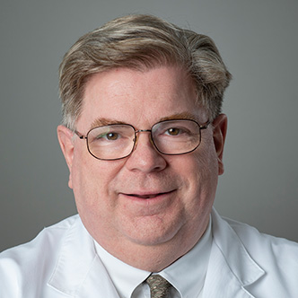 Dr. Cotterell has short blond hair, is wearing glasses and a dress shirt and tie under a white medical coat, and is smiling at the camera