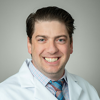 Dr. DeBiase has short brown hair, is wearing a plaid shirt with blue and orange lines, a matching tie under a white medical coat, and is smiling at the camera.