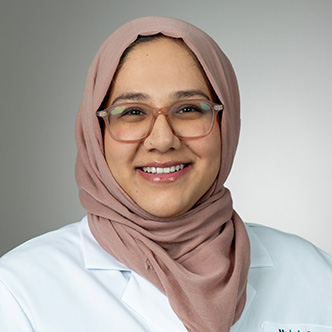Dr. Madeeha is wearing a light mauve head covering and white medical coat. She has glasses and is smiling at the camera.