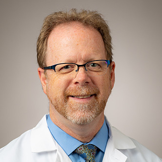 Dr. Theisz has short red hair and graying goatee, wears glasses, and is wearing a blue shirt and tie under a white medical coate