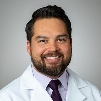 Dr. Gordon has short dark hair and goatee, is wearing a light dress shirt and dark tie under a white medical coat, and is smiling at the camera
