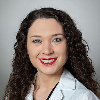 Anna Louthan has long, dark curly hair, is wearing a white medical coat, and is smiling at the camera.