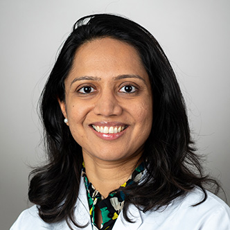 Dr. Shah has long dark hair, is wearing a white medical coat and is smiling at the camera