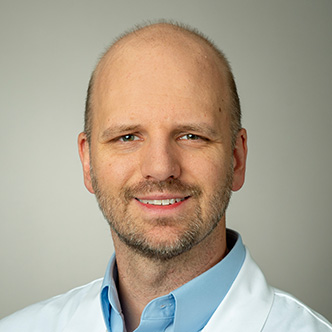 Dr. Sheets has very short hair, a beard, is wearing a blue dress shirt under a white medical coat, and is smiling at the camera.