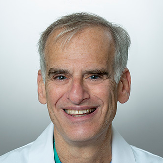 Dr. Spiegel has short grey hair, is wearing blue scrubs and a white medical coat, and is smiling at the camera