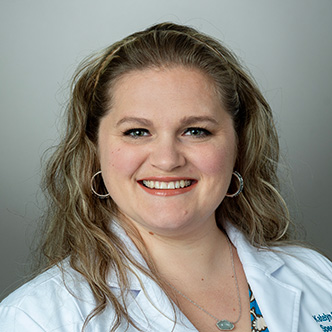 Katelyn has long and wavy dark blond hair, is wearing a white medical coat and is smiling at the camera