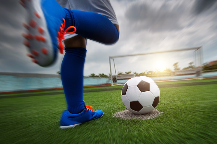 Close up of legs of a person kicking a soccer ball. The lighting is dramatic and the person is wearing bright blue gear and shoes with orange laces.