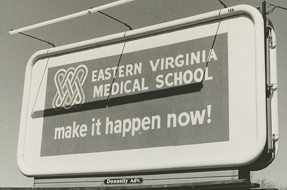 A grayscale photograph from the 1960s showing a billboard with the words Eastern Virginia Medical School make it happen!