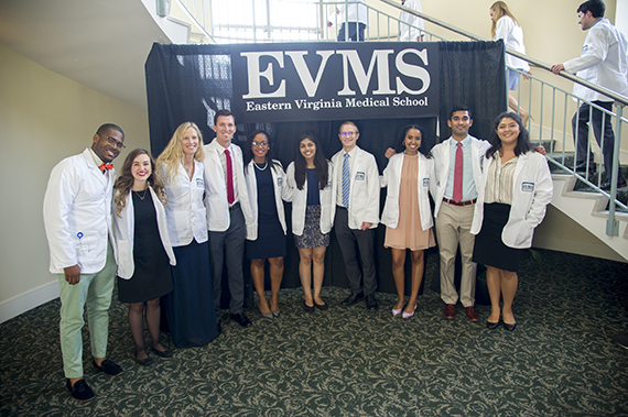 10 students, male and female, stand in front of an EVMS sign wearing their short EVMS white coats 