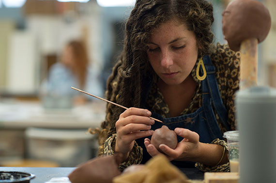 A young woman with long brown hair uses a tool to shape a clay figure. A head shaped clay figure is in the foreground.