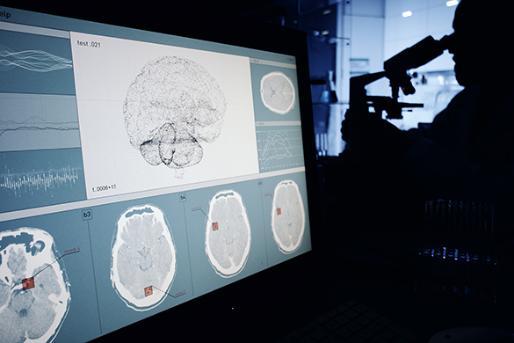 Several images of brainwave scans are shown on a screen in the foreground while a man looks at a microscope in the background
