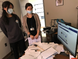 Two masked women standing next to a messy desk holding a computer monitor