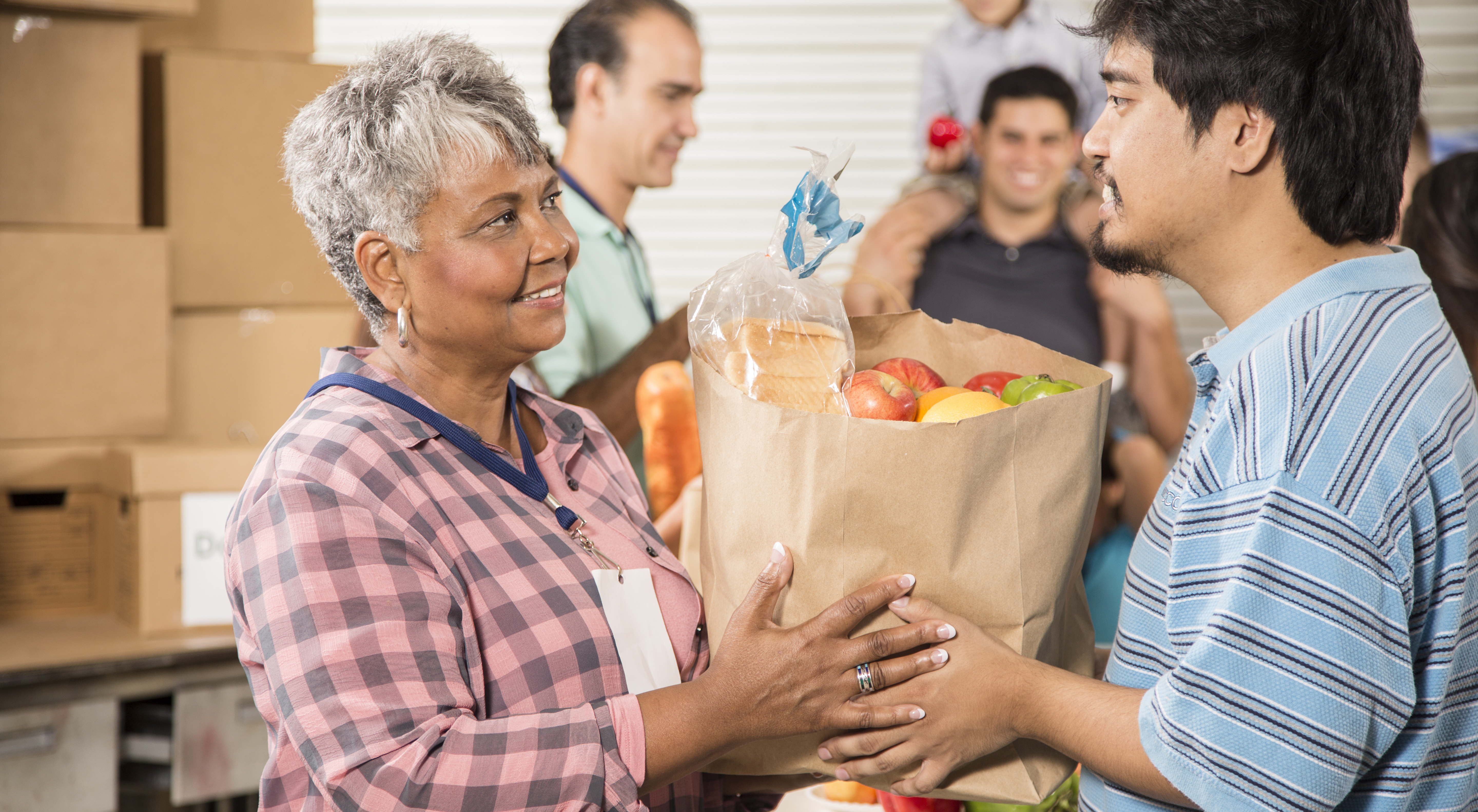 A food pantry employee hands a bag of food to another person, with several volunteer and clients in the background.