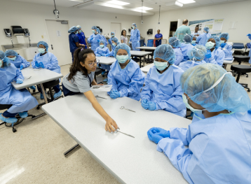 students and instructors wearing blue scrubs, gloves, and hair nets in classroom. 