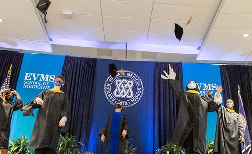 Five masked grads toss their caps on a stage before the EVMS seal