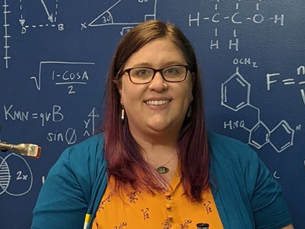Woman with brown hair in yellow blouse and teal sweater smiles while standing in front of science chalkboard