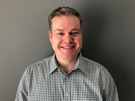 A man with short hair in a plaid shirt smiles at the camera. There is a dark gray wall behind him.