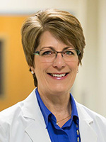 A woman with light brown hair and glasses wearing white lab coat smiles at the camera