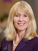 A woman with blonde hair wearing a purple jacket smiles at the camera