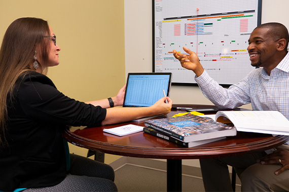 A student sitting at a table with a professor working on a research project. There's a bulletin board behind them with a timeline of dates.