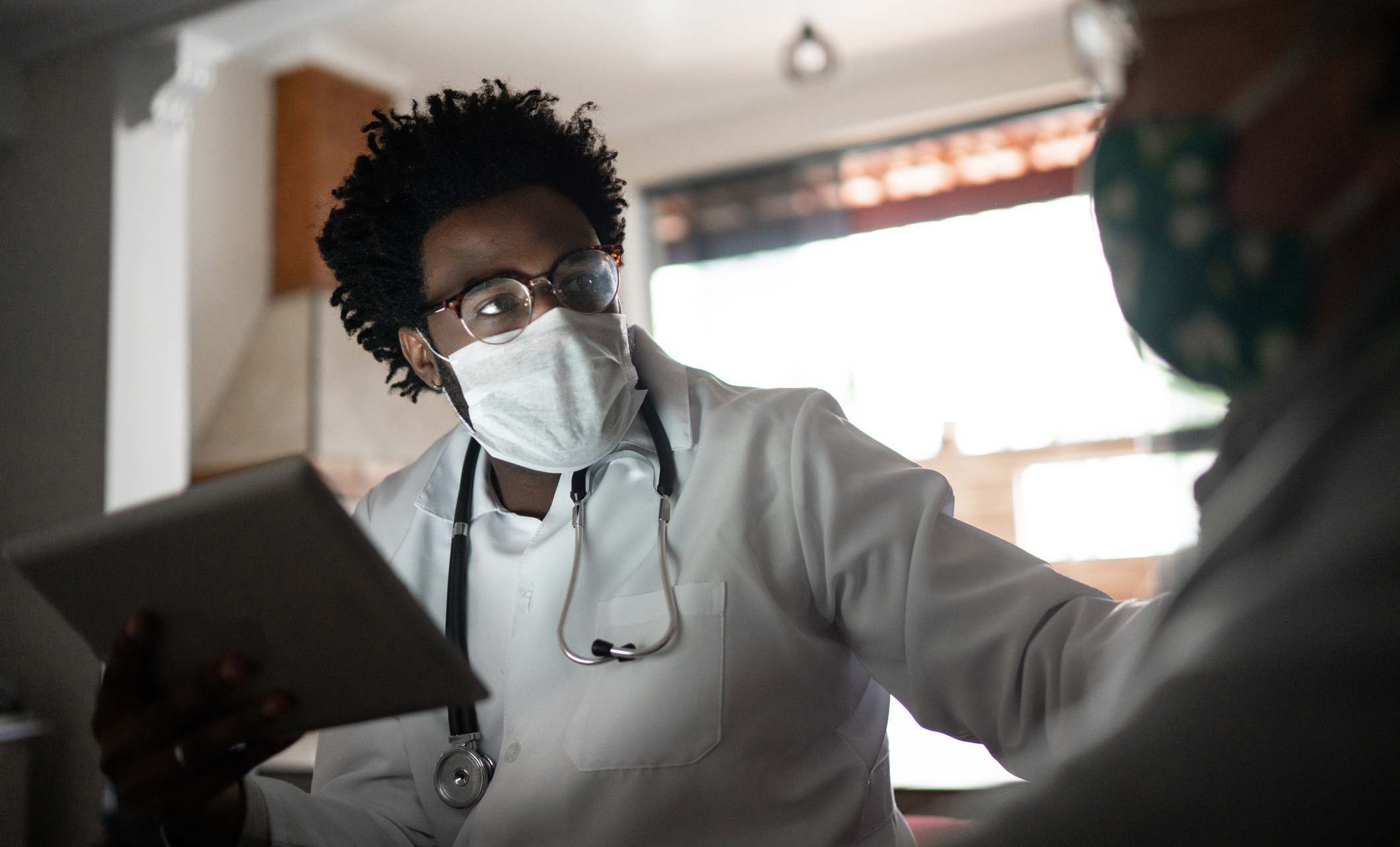 A younger physician assistant wearing glasses, white coat and stethoscope, examines patient while holding digital pad