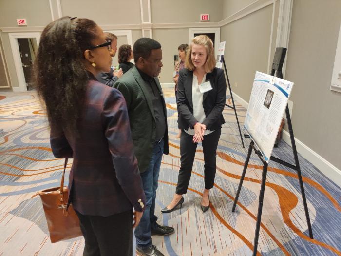 EVMS Medical Student Irene Peppiatt is standing with her poster presentation, presenting it to two event participants. They are standing in a conference room environment.