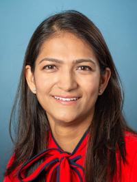 Dr. Patel has long, straight dark hair, is wearing a bright red blouse with a bow around the neck, and is smiling at the camera.