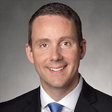 headshot of smiling man in a dark suit, white shirt and blue tie