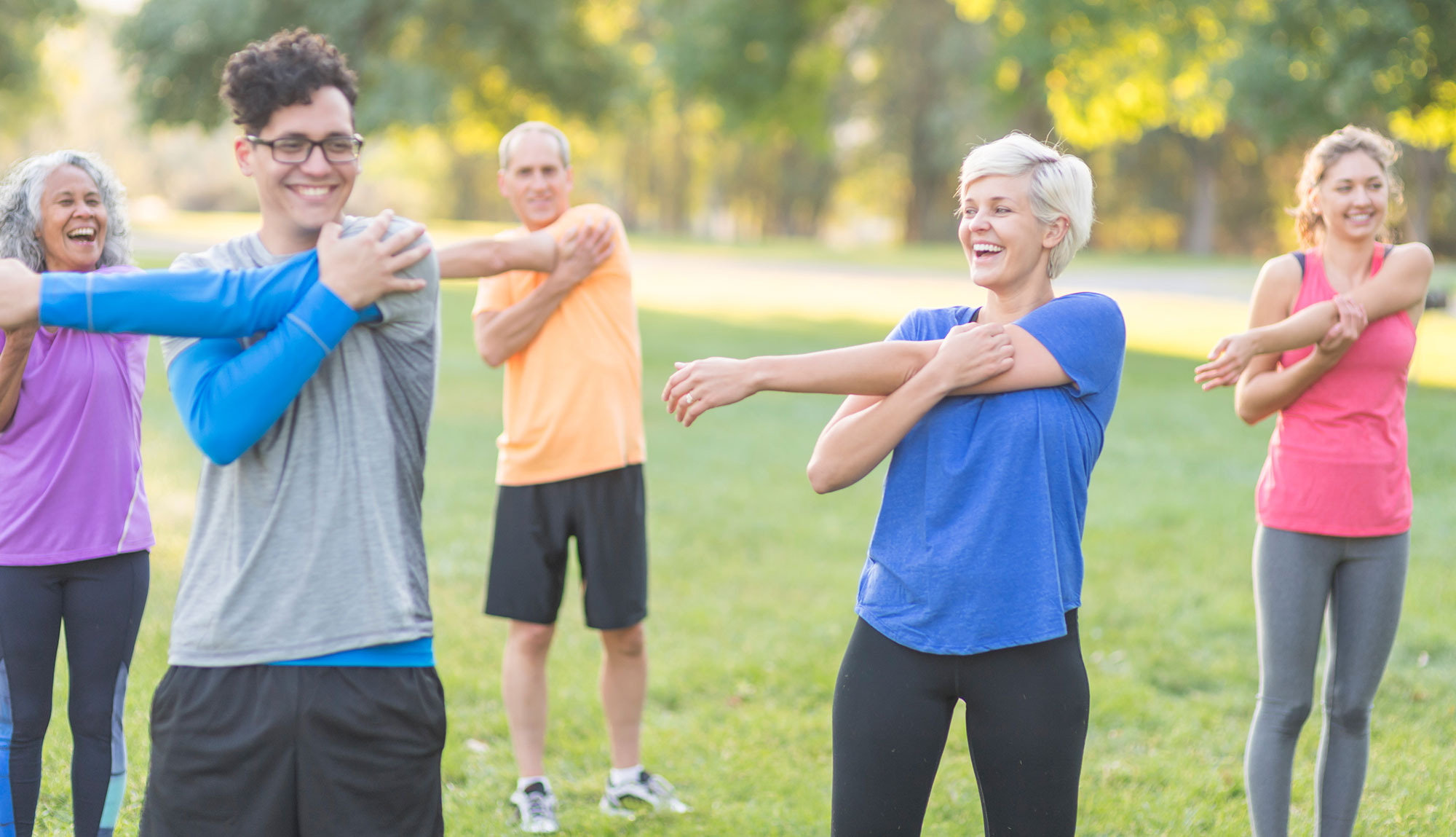 A fitness group of five people stretch their arms together while smiling and laughing on the lawn of a park.