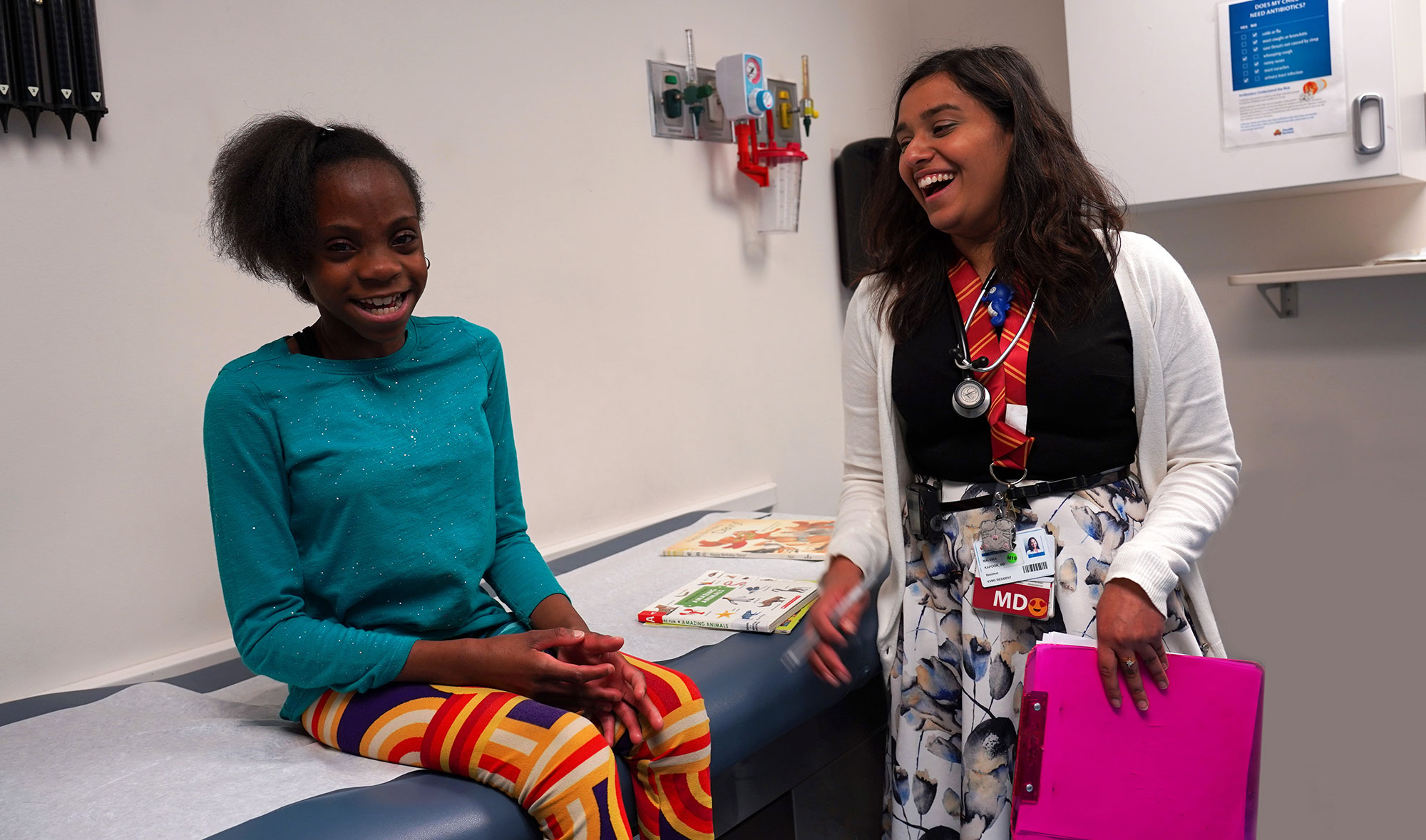 Dr. Kapoor laughs with a young girl, who is smiling while waiting for her checkup, in an exam room.