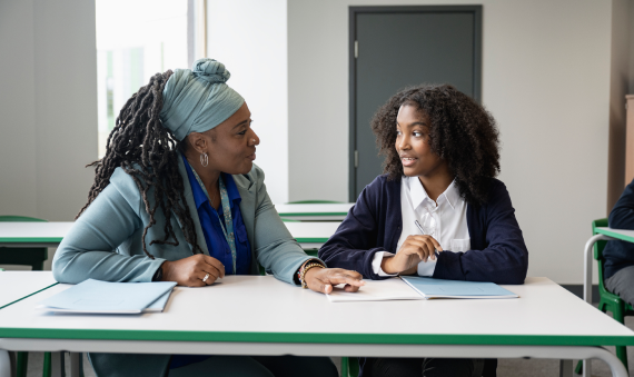 Supportive female teacher sitting at desk with teenage schoolgirl