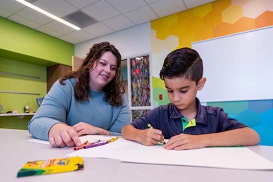 A female therapist in blue shirt sits with a boy as he colors with crayons. They are in a colorful room.