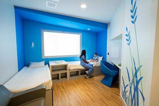 A female therapist in blue medical scrubs sits on a bed in a patient's room speaking to a child.