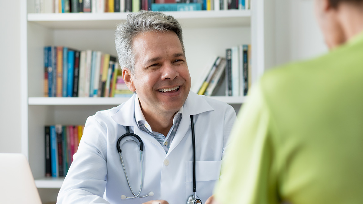 A male doctor with short gray hair wearing a white medical coat and stethoscope smiles at a patient while sitting in an office.