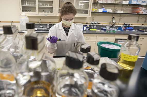 A student where a PPE mask, lab coat and gloves works in a research lab.