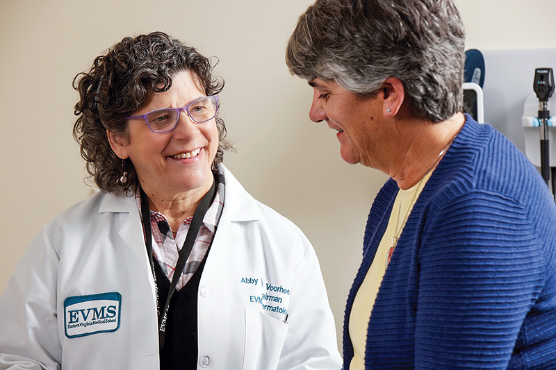 Dr. Abby Van Voorhees has medium curly hair, is wearing glasses and a stethoscope around her neck, a white medical coat, and is smiling at a patient, an older woman with graying hair.