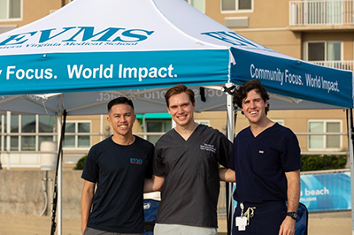 Three men wearing dark surgical scrubs smile at the camera while standing next to a pop-up tent on the beach